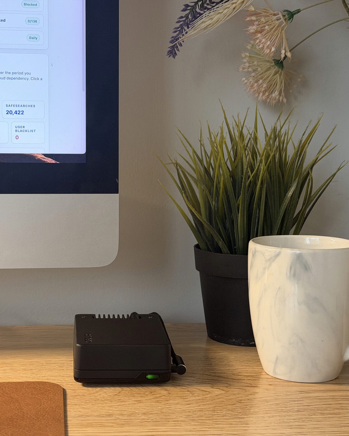 Shield Core on an oak desk beside a plant and a mug, green status light on, with part of the Shield dashboard on the iMac behind it.