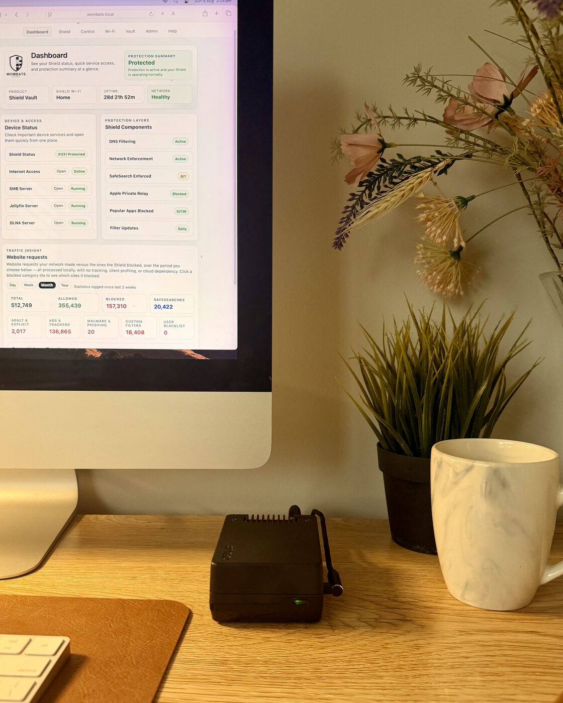 Shield Vault on an oak desk beside a plant and a mug, green status light on, with its dashboard on the iMac behind it showing Protected and 28 days of uptime.
